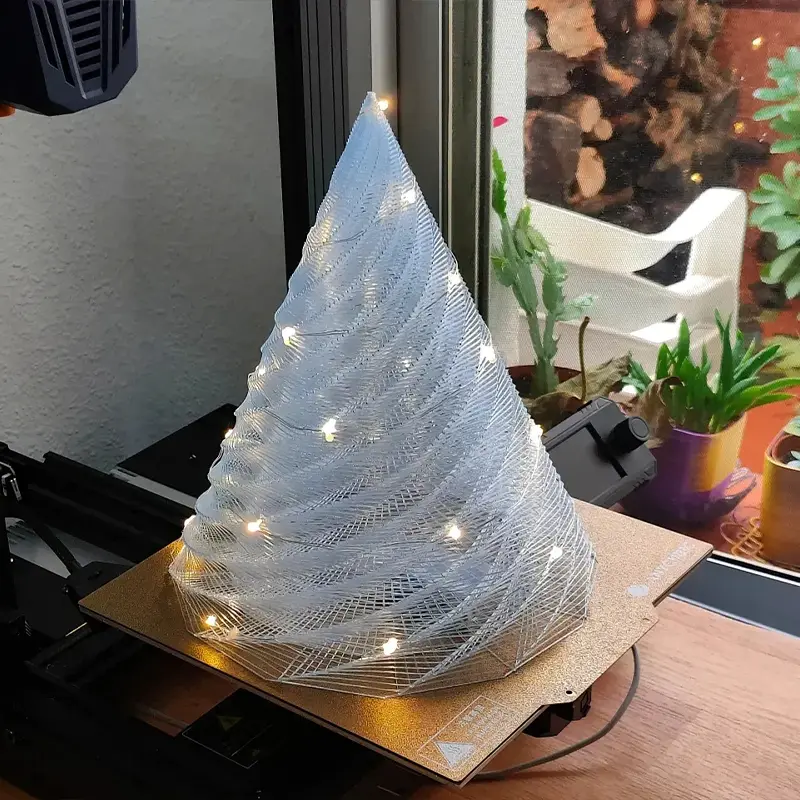 Silver-gray 3D printed spiral Christmas tree with warm white LED lights glowing on wooden table in modern home setting.