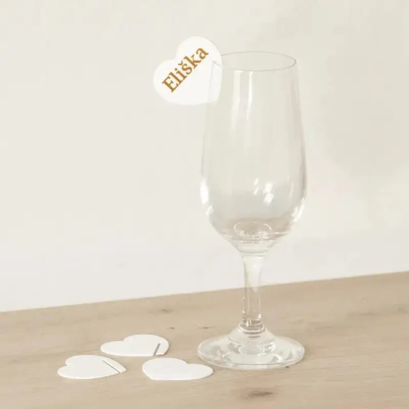 A wine glass with a white heart-shaped ornament bearing the name "Eliška", set on a wooden background with additional ornaments