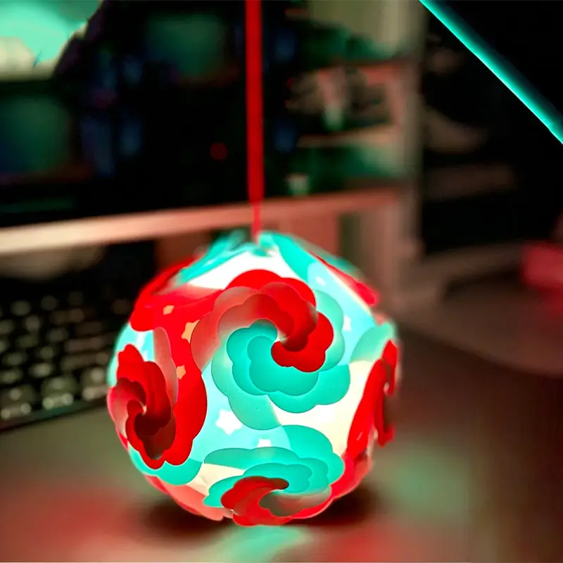 A detailed view of a bespoke 3D printed illuminated orb featuring intricate red and teal swirling patterns, hanging against a blurred workspace background with keyboard and monitor.