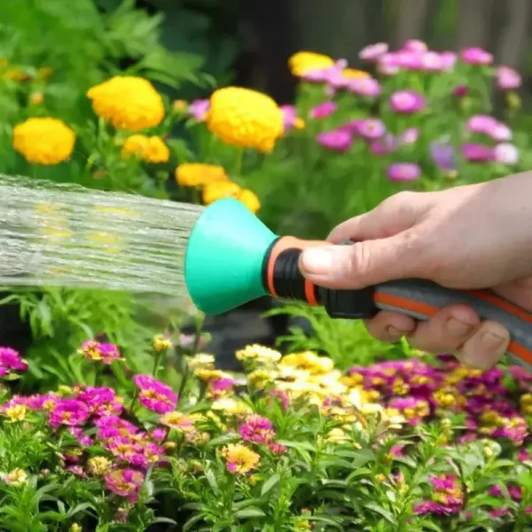 Hand holding a green precision garden spray nozzle emitting fine water mist, with colorful flowers in the background