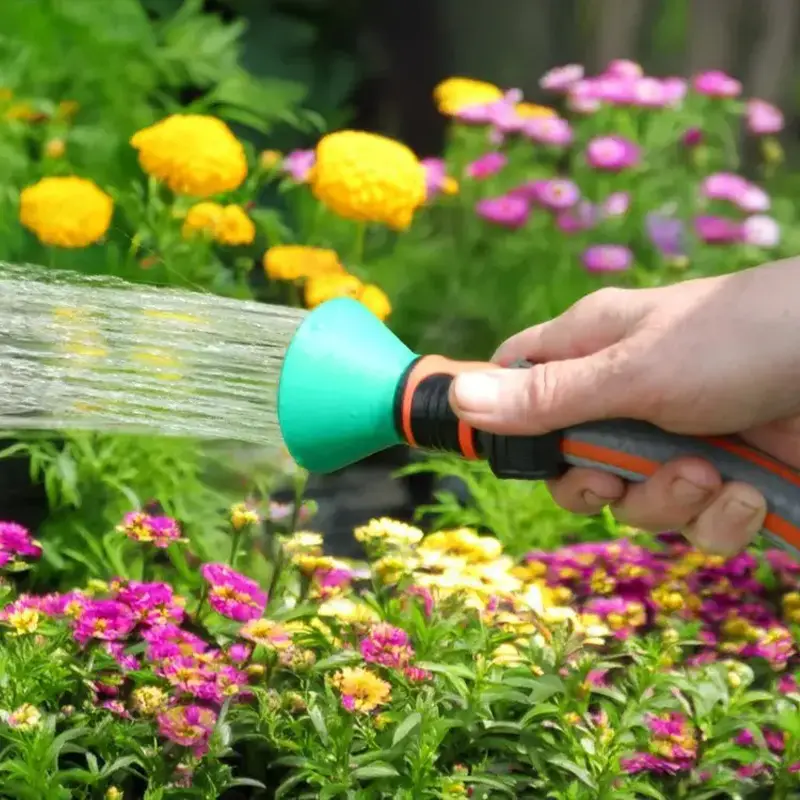 Hand holding a green precision garden spray nozzle emitting fine water mist, with colorful flowers in the background