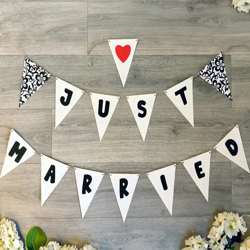 White triangular bunting spelling "JUST MARRIED" with black damask patterns and a red heart, displayed on a wooden floor