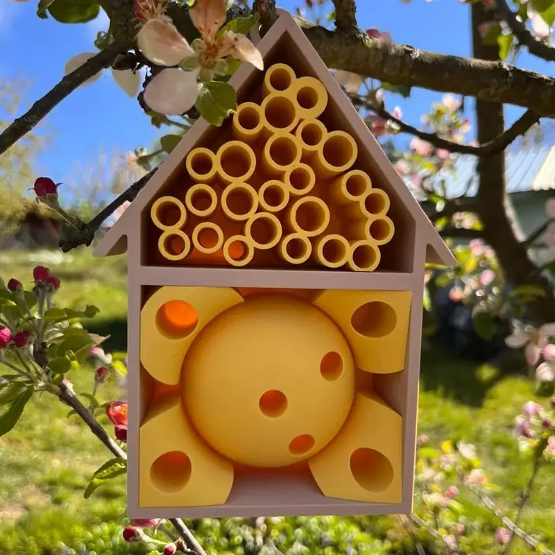 Custom 3D printed insect hotel hanging from a tree branch amidst blooming flowers, showcasing eco-friendly design for B2B clients