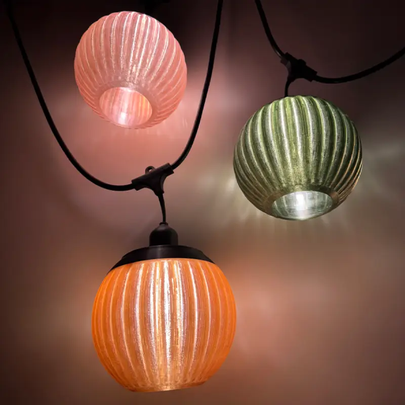 Three colorful 3D printed bulb shades in pink, green, and orange hanging from black cords, showcasing ribbed texture and soft glow against plain background