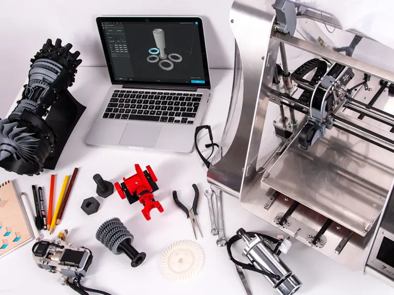A professional 3D printing workspace featuring a silver 3D printer, open laptop displaying 3D models, and various custom-printed mechanical parts in black and red colors, arranged with tools on a clean white desktop background for B2B clients.