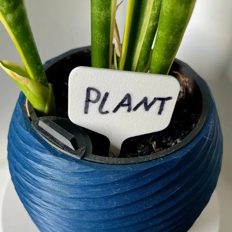 White 3D printed botanical identification tag reading "PLANT" inserted in blue vase with green plants