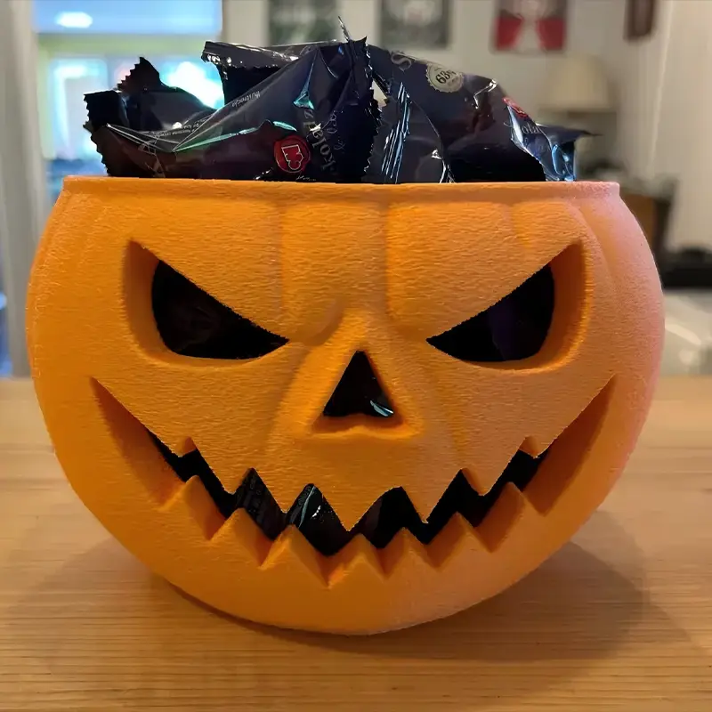 Orange 3D printed pumpkin-shaped bowl with a scary face, filled with black-wrapped chocolates for Halloween events and B2B clients